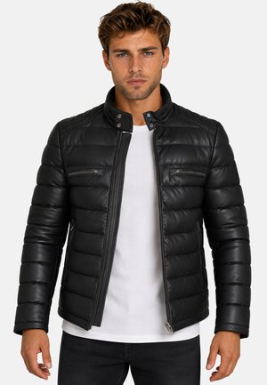 Hombre vestido con una chaqueta de cuero negra acolchada sobre una camisa blanca, de pie frente a un fondo liso de color gris claro.