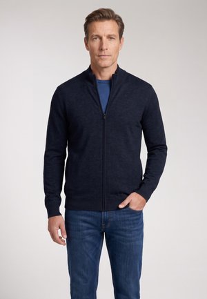 Mann steht nach vorne gewandt und trägt einen marineblauen Reißverschluss-Pullover über einem blauen Hemd und blauen Jeans, mit einer Hand in der Tasche.