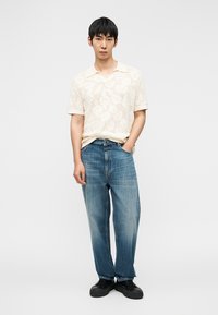 Μπεζ κοντομάνικο πόλο πουκάμισο με λουλουδάτο σχέδιο, συνδυασμένο με ανοιχτό μπλε φαρδιά τζιν και μαύρα αθλητικά παπούτσια, σε χονδροειδές φόντο.