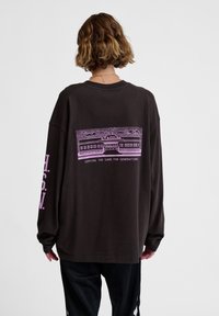 Dunkelbraunes Langarmhemd mit einem rosa Grafikdesign eines Stadions und Text, mit lockerer Passform und weichem Baumwollmaterial.