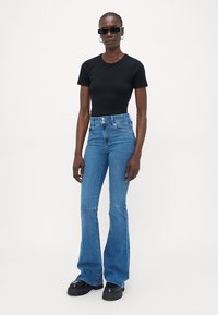 Mujer vestida con una camiseta ajustada negra de manga corta, jeans de tiro alto azules de campana, zapatos negros gruesos, pendientes de aro y gafas de sol negras estrechas.