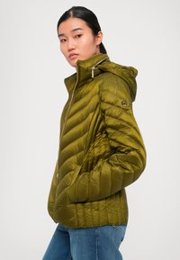 Kvinne med mørkt hår samlet i hestehale, iført en olivengrønn quiltet jakke med hette og blå jeans, ser sidelengs med hendene i lommene.