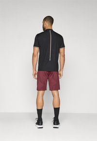 Polo atlético negro con cremallera en la espalda y acentos blancos, combinado con pantalones cortos burdeos que presentan un patrón geométrico texturizado. Zapatillas y calcetines negros.