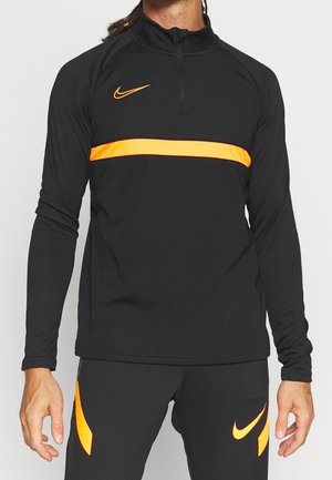 Homme portant un survêtement Nike noir avec une bande orange sur la poitrine et des accents orange sur le pantalon, debout devant un fond uni.