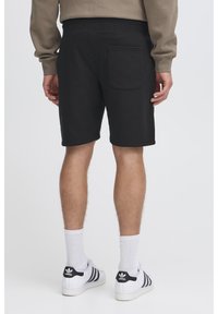 Shorts en coton noir au design simple, avec une poche arrière et une taille élastique. Portés avec des baskets blanches et noires.