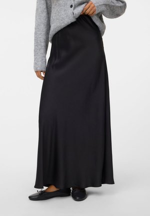 Mujer con una falda larga de satén negra, zapatillas de ballet negras y un suéter de punto gris, sujetando ligeramente el suéter hacia arriba.