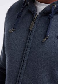 Primo piano di una persona che indossa una felpa con zip blu scuro con cordini e una maglietta bianca sotto.