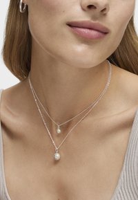 Collares de plata en capas con un pequeño colgante redondo que tiene una perla y un colgante de gema más pequeña, descansando sobre un escote neutro.