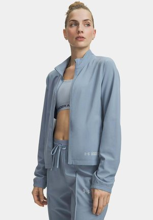 Mujer que lleva chaqueta deportiva Under Armour azul claro, sujetador deportivo y pantalones con cordón, mirando hacia un lado sobre un fondo liso.