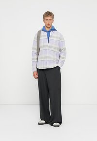 Camisa de botones con diseño de cuadro lila y blanco, superpuesta a una sudadera azul. Pantalones anchos negros y zapatillas gris/blancas.