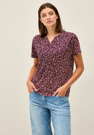 Chemise à manches courtes de couleur violette avec un motif de feuilles, dotée d'un col en V et d'un tissu doux. Assortie avec un jean en denim bleu clair.