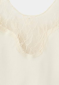 Tissu couleur crème avec une fine superposition de dentelle florale au niveau de l'encolure, présentant des motifs délicats et complexes de dentelle ainsi qu'une texture textile lisse.