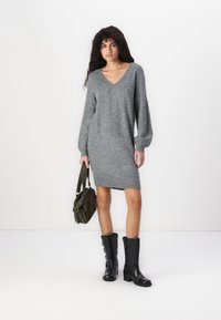 Robe grise en tricot avec un décolleté en V, de longues manches ballon et une coupe droite, associée à des bottes noires jusqu'aux genoux et un sac à bandoulière vert.