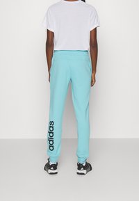 Person som har på sig ljusblå Adidas byxor med svart logotyp på vänster ben, vit t-shirt och svarta Adidas sneakers, står vänd bort.