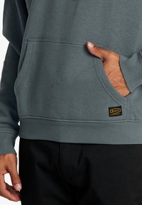 Sudadera gris con un bolsillo tipo canguro, dobladillo y puños acanalados. Presenta una etiqueta con el logo de RVCA en negro y amarillo en el lado izquierdo del frente.