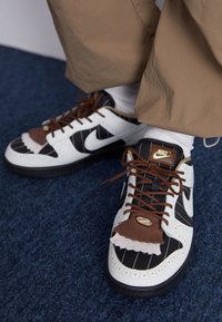 Nike-tennarit, joissa valkoinen nahkainen ja musta raidallinen muotoilu. Ruskeat korostukset varpaan kohdalla, teksturoidut yksityiskohdat ja kultainen logokyltti sivulla.