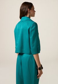 Giacca corta color teal con colletto strutturato e maniche a tre quarti, abbinata a pantaloni coordinati. Tessuto liscio con un design moderno.