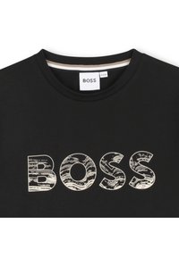 Melnā T-kreklā ar apaļu kakla izgriezumu, uz kura redzams vārds "BOSS" tekstūrētā krēmkrāsas rakstā. Etiķete redzama iekšpusē ap kaklu.