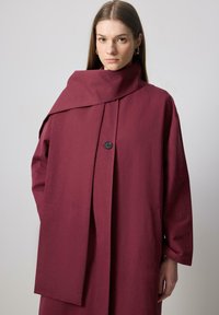 Burgundský oversize kabát s vysokým golierom, so zapínaním na gombík a dvoma prednými vreckami. Hladká tkanina s minimalistickým dizajnom.