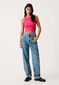 Aansprechende roze mouwloze crop top met logo, gecombineerd met hoge taille, loszittende blauwe spijkerbroek en zwarte instappers.