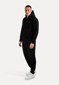 Sudadera negra con cremallera y bolsillo tipo canguro, combinada con pantalones de chándal negros a juego y zapatillas blancas. Textura suave, diseño simple.