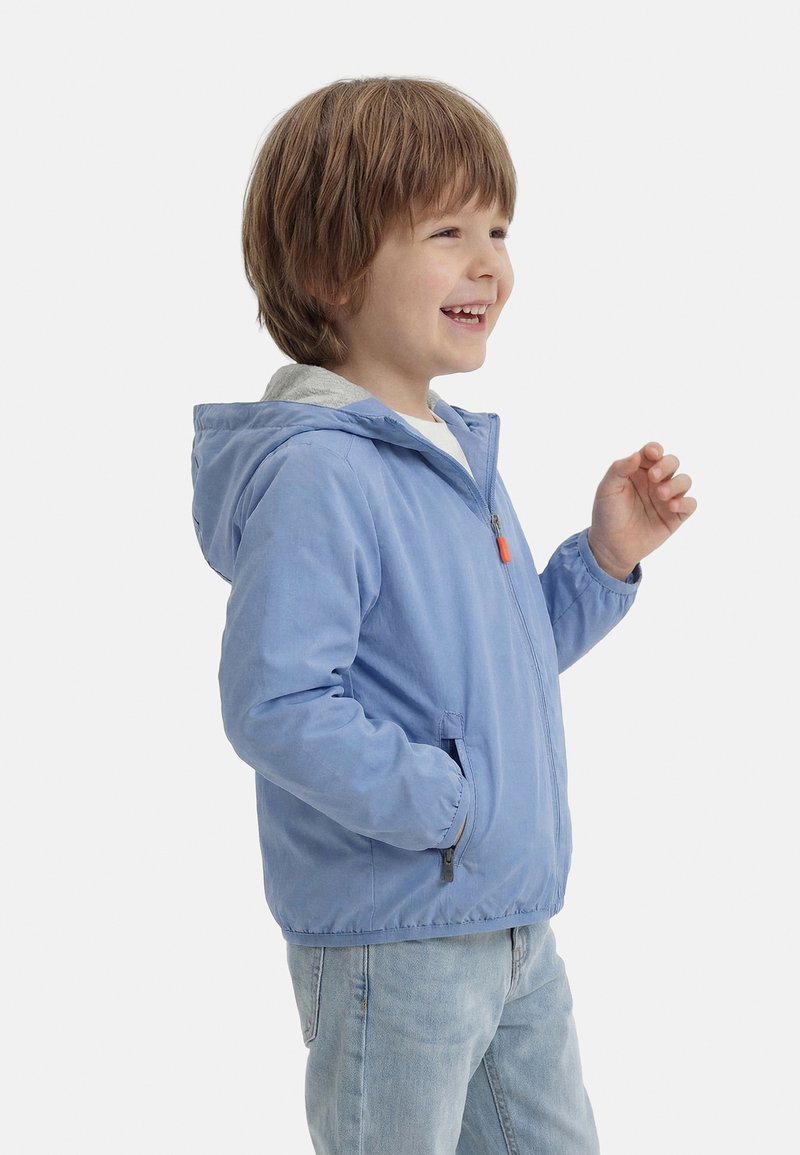 Νεαρός αγόρι που χαμογελά, φοράει ένα γαλάζιο μπουφάν με κουκούλα και τζιν σε ανοιχτό ξέβαμμα, στέκεται μπροστά σε ένα απλό λευκό φόντο.