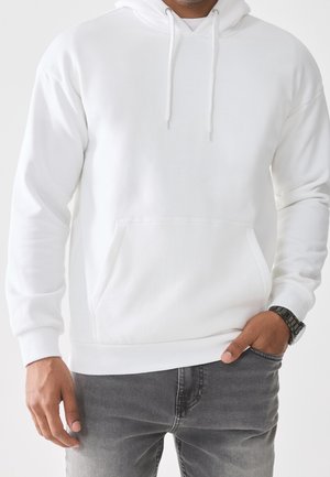 Mand iført enkel hvid hættetrøje med forlomme og grå jeans, hånd i lommen og sort armbåndsur på venstre håndled.