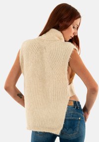 Suéter sin mangas de punto en suave hilo color crema, con un alto cuello, textura acanalada y lados abiertos para un ajuste relajado.