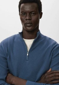 Modra rebrasta jopica na zadrgo z visokim ovratnikom, nošena čez kremasto srajco. Model stoji s prečkanimi rokami, kar prikazuje teksturo in prileganje oblačila.