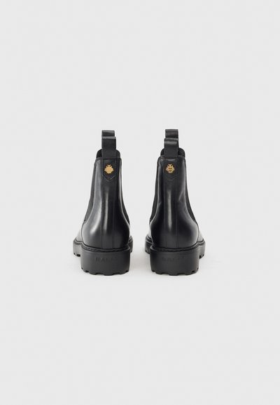 Botine Chelsea din piele neagră, cu tălpi groase, panouri elastice laterale și bucle de tragere. Accentele de hardware aurie prezintă un logo pe toc.