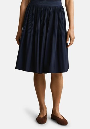 Femme portant une jupe midi plissée bleu marine et des chaussures plates marron, debout avec les mains détendues le long du corps sur un fond blanc.