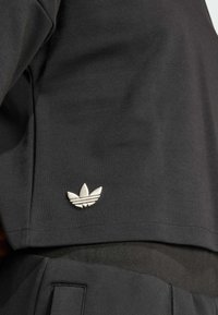 Čierne bavlnené tričko s krátkymi rukávmi, s bielou vyšívanou logom troch listov na spodnej strane. Hladká textura, voľný strih.