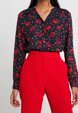 Frau trägt eine marineblaue Bluse mit roten Blumen- und orangefarbenen Punktmustern, die in eine hochtaillierte, leuchtend rote Hose gesteckt ist.