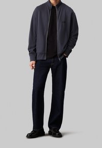 Zip-up sweatshirt i mörkgrå, texturerat tyg; kombinerad med en svart tröja, mörkblå jeans och svarta skor med låg profil.