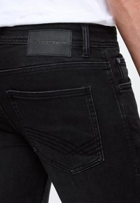 Fekete farmernadrág, márkajellel ellátott bőrpatkóval, látható varrással és ívelt kialakítású hátsó zsebbel.