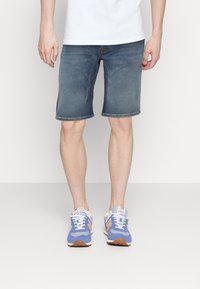 Shorts de mezclilla en azul medio con corte recto, con bolsillos delanteros y cremallera. Combinados con zapatillas deportivas azul y naranja.
