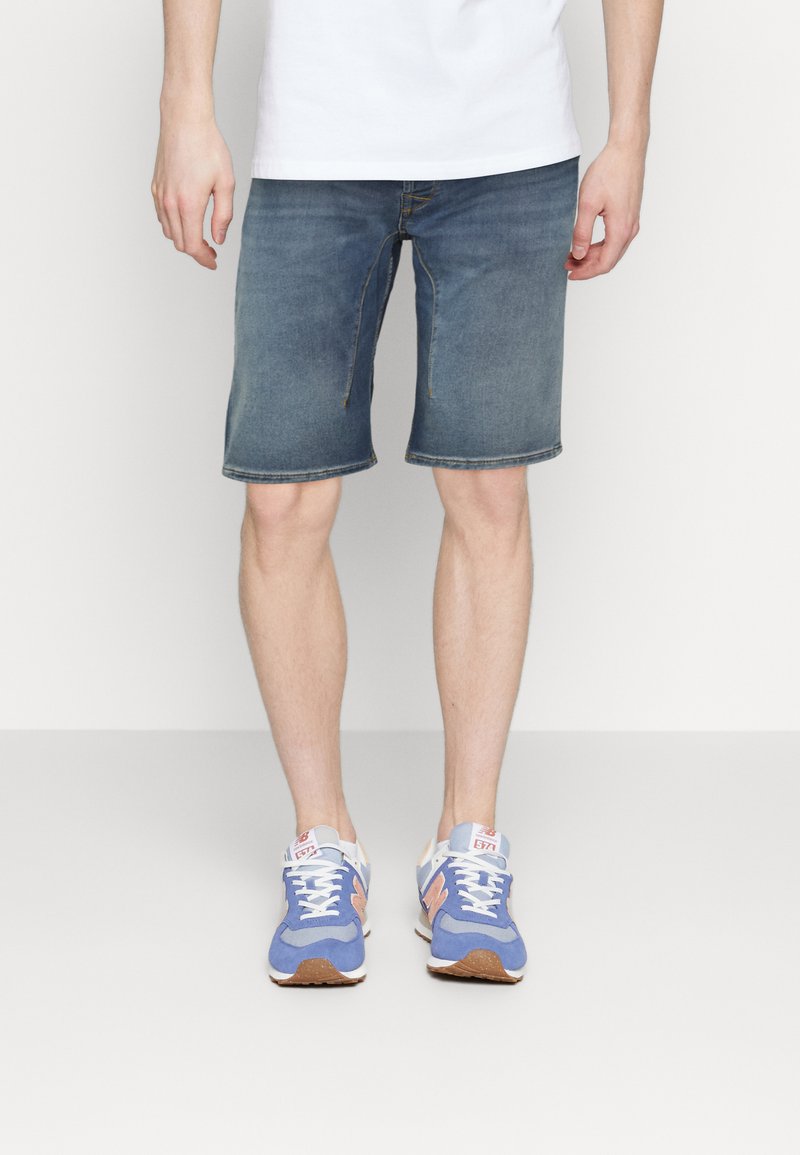 Shorts de mezclilla en azul medio con corte recto, con bolsillos delanteros y cremallera. Combinados con zapatillas deportivas azul y naranja.