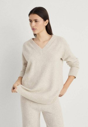 Pull v-neck beige en tissu doux et texturé, avec des poignets et un ourlet côtelés, porté avec un pantalon beige clair assorti.