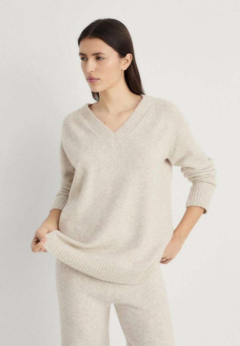 Beige V-Ausschnitt-Pullover aus weichem, strukturiertem Stoff mit gerippten Bündchen und Saum, getragen mit passenden hellbeigen Hosen.