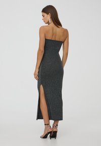 Abito midi nero senza spalline con una texture luccicante, silhouette aderente e spacco laterale. Abbinato a décolleté nere con cinturini.