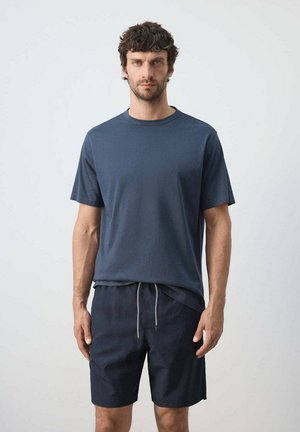 Homme aux cheveux bouclés et foncés, debout face à l'avant, portant une chemise à manches courtes bleu marine et un short foncé avec cordon, sur un fond uni.