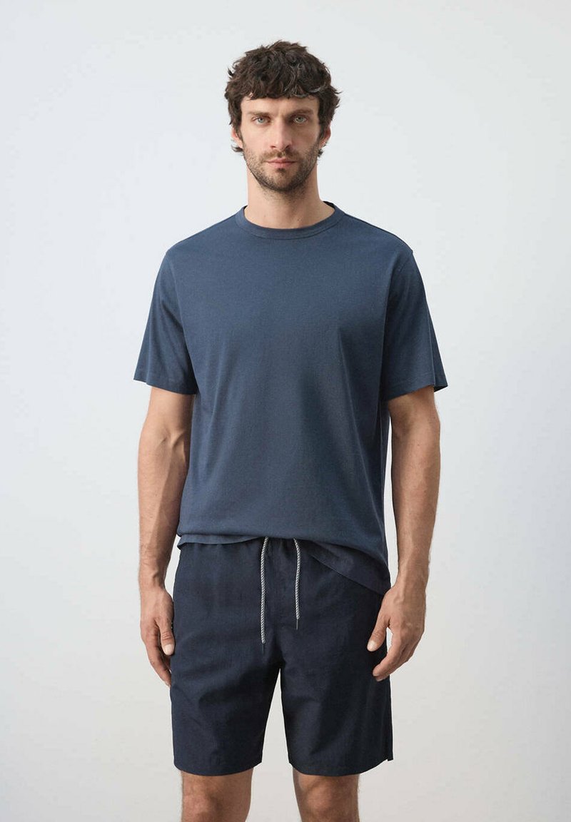 Homme aux cheveux bouclés et foncés, debout face à l'avant, portant une chemise à manches courtes bleu marine et un short foncé avec cordon, sur un fond uni.