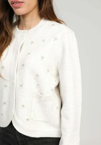 Femme portant un cardigan blanc à boutons avec de petites embellissements florales en perles et fil, ainsi qu'une poche à bouton sur le devant.