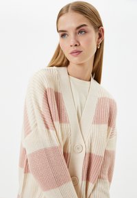 Stickad kofta med rosa och krämfärgade ränder, med en djup V-ringning, två knappar och strukturert tyg. Avslappnad design med långa ärmar.