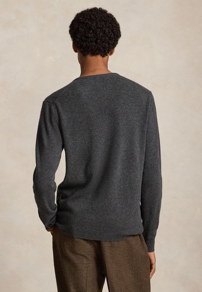 Uomo con capelli ricci che indossa un maglione a maniche lunghe grigio scuro e pantaloni marroni a quadri, in piedi di fronte a una parete beige.