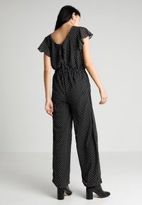 Combinaison noire à pois avec manches courtes à volants, taille élastique, jambes amples, et accents noirs unis. Bottines noires à talons hauts.