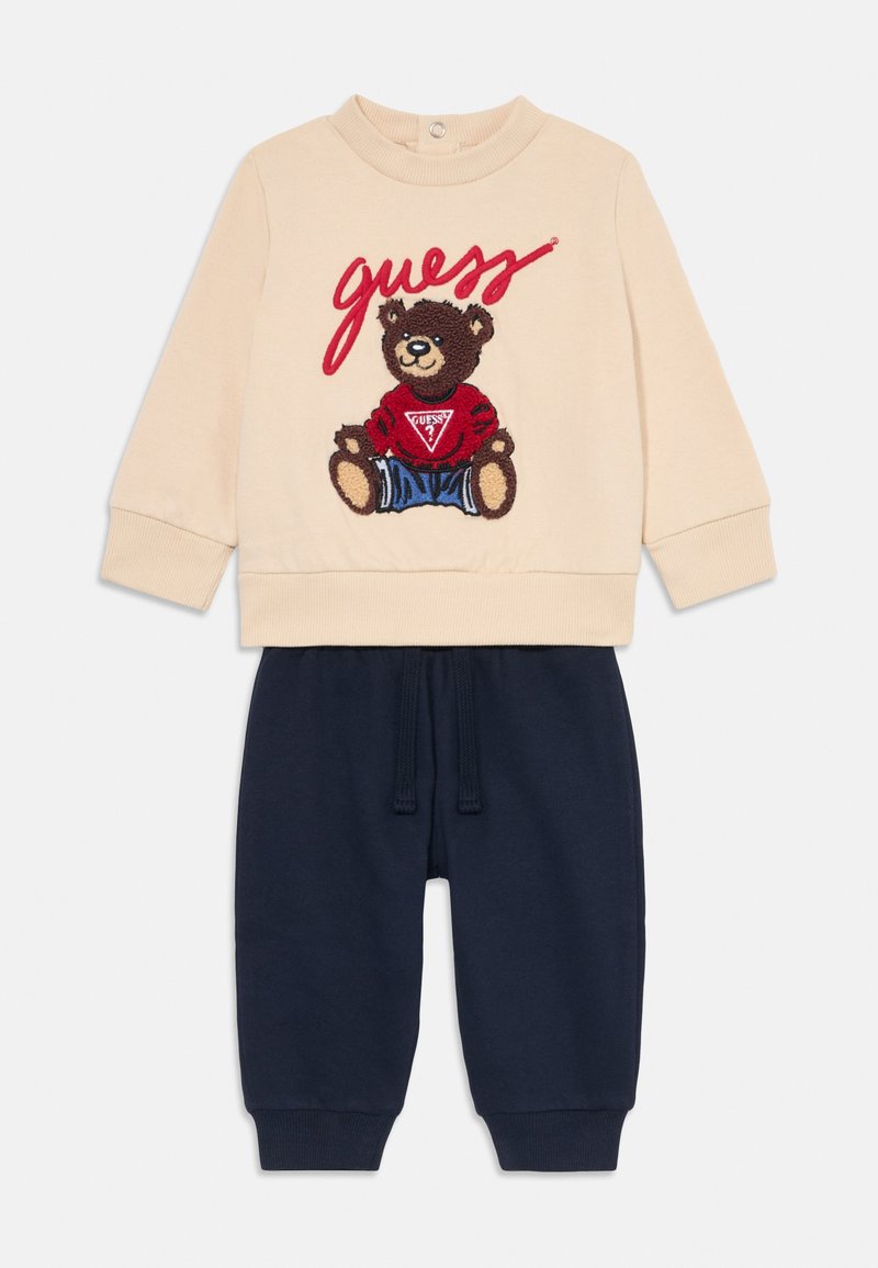 Beige hettegenser med bjørnetrykk og "guess"-tekst; marineblå joggebukser med elastisk midje og mansjetter. Blandingsstoff av bomull.