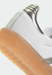 Białe sneakersy z gładką skórzaną cholewką, metalicznym srebrnym akcentem na pięcie oraz przezroczystą gumową podeszwą z teksturowanym wzorem.