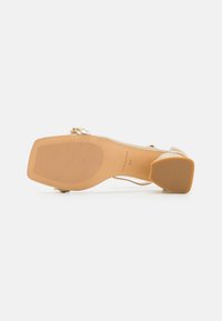 Sandal de tacón alto beige con parte superior de cuero suave, punta cuadrada y suela de goma texturizada. Presenta acentos en tono dorado y tiras delgadas.