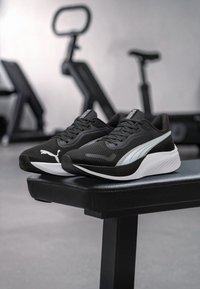 Schwarze Sportschuhe mit einem strukturierten Mesh-Obermaterial, glatten weißen Akzenten und einer gepolsterten Sohle, die auf einer schwarzen Bank positioniert sind.